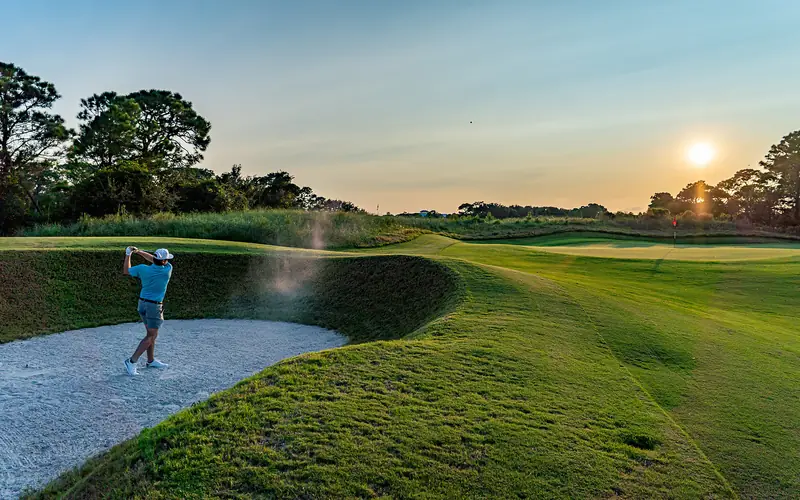 Golfer hitting out of a greenside bunker at sunset